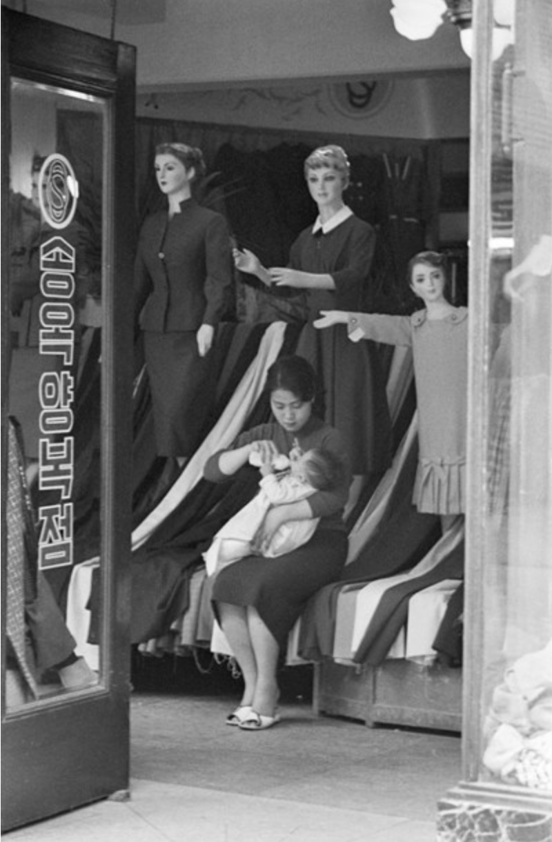 1950년대 서울의 한 양복점에서 여성이 아이에게 젖병을 물리고 있다. 핑크칼라에게 가정과 일은 선택이 아니라 융합으로 새롭게 창조했던 그들만의 고유한 영역이었을지 모른다. 사진=한영수문화재단.